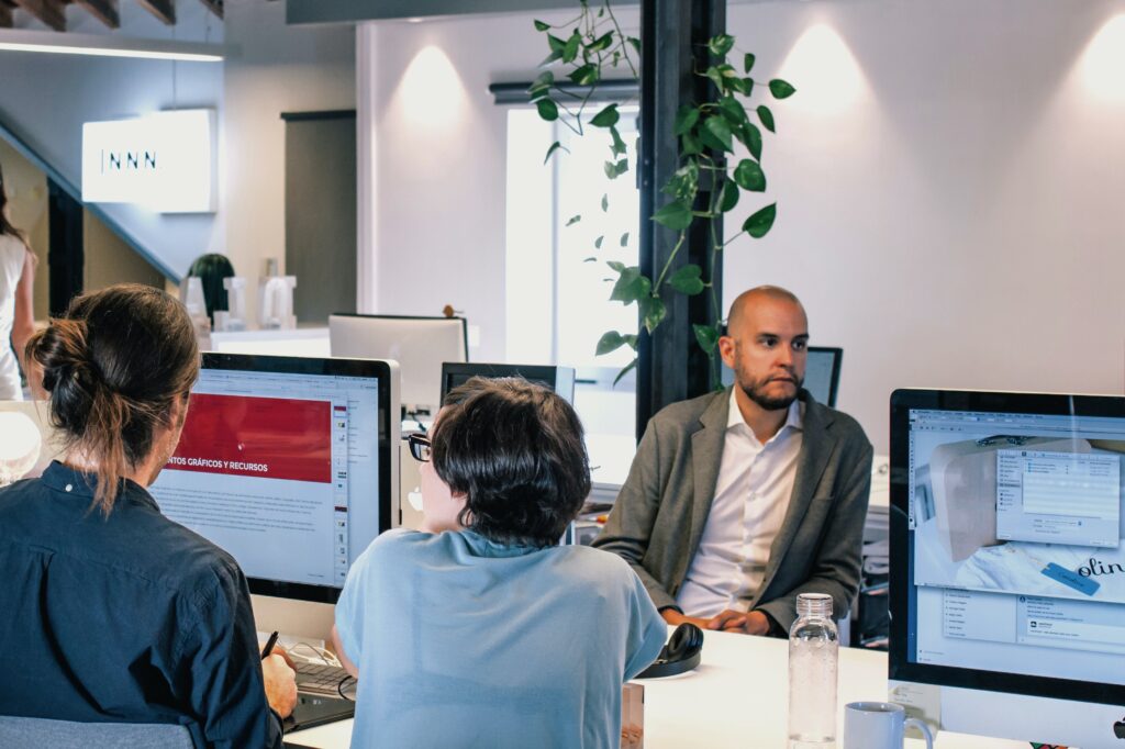 Image of three people in an open office plan. Two people have their backs turned looking at one Apple computer screen. One person is looking off in the distance with a negative facial expression.