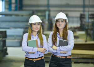 An image of two women in construction. They are wearing purple collared shirts, black pants, and white hard hats.